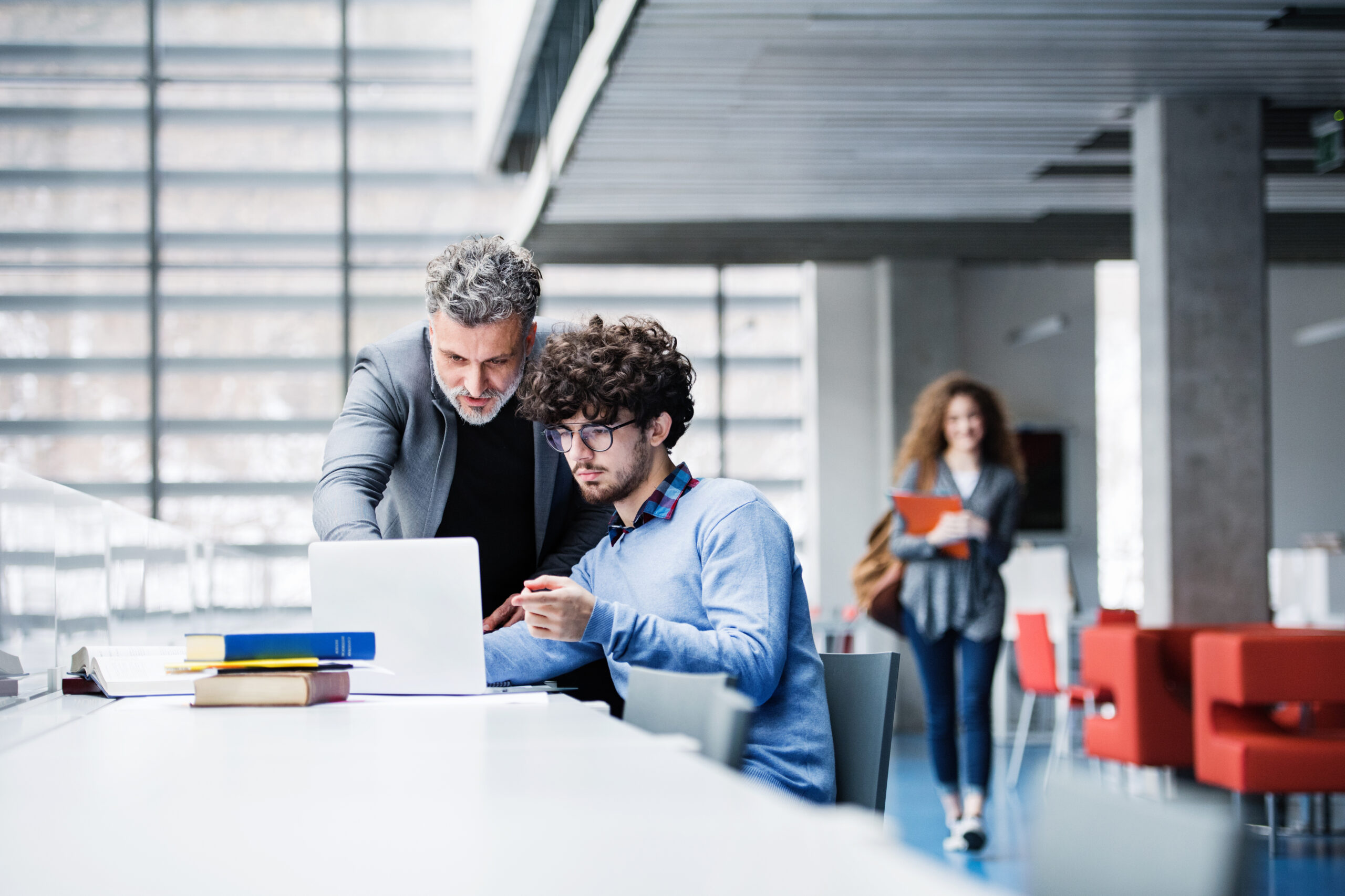 Handsome university professor helping student with final project, showing him research paper, book for thesis in library.